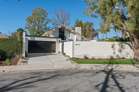 White home behind a low stucco wall, with a dark garage door and mature trees, seen from the street